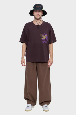 Mężczyzna w brązowym oversized t-shircie z nadrukiem, szerokich spodniach i ciemnym kapeluszu typu bucket hat, styl streetwear