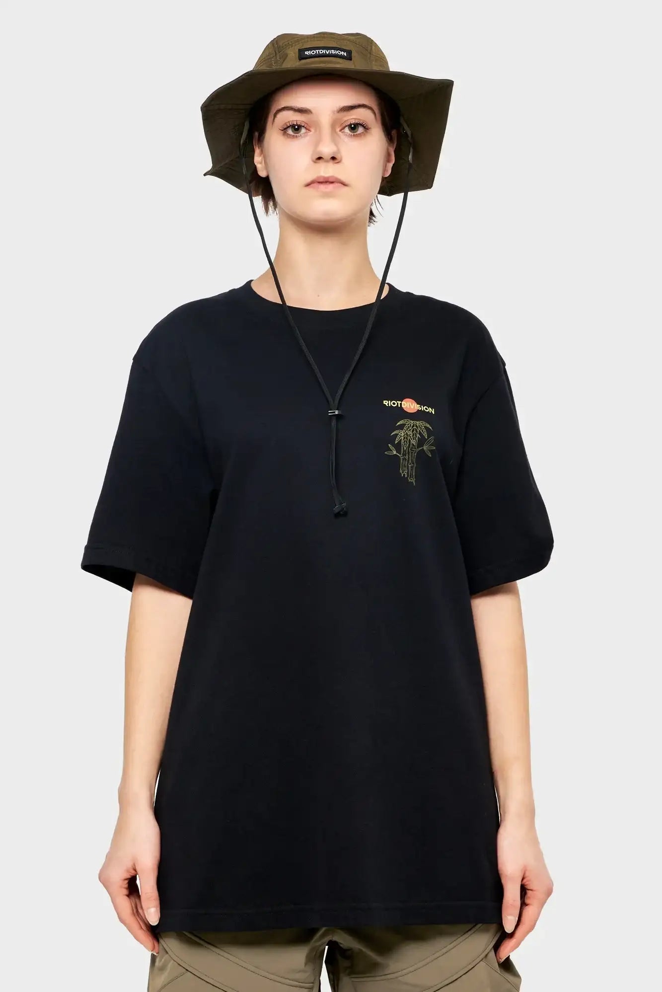 Osoba w czarnym t-shircie RIOTDIVISION z nadrukiem i oliwkowym kapeluszu, styl streetwear