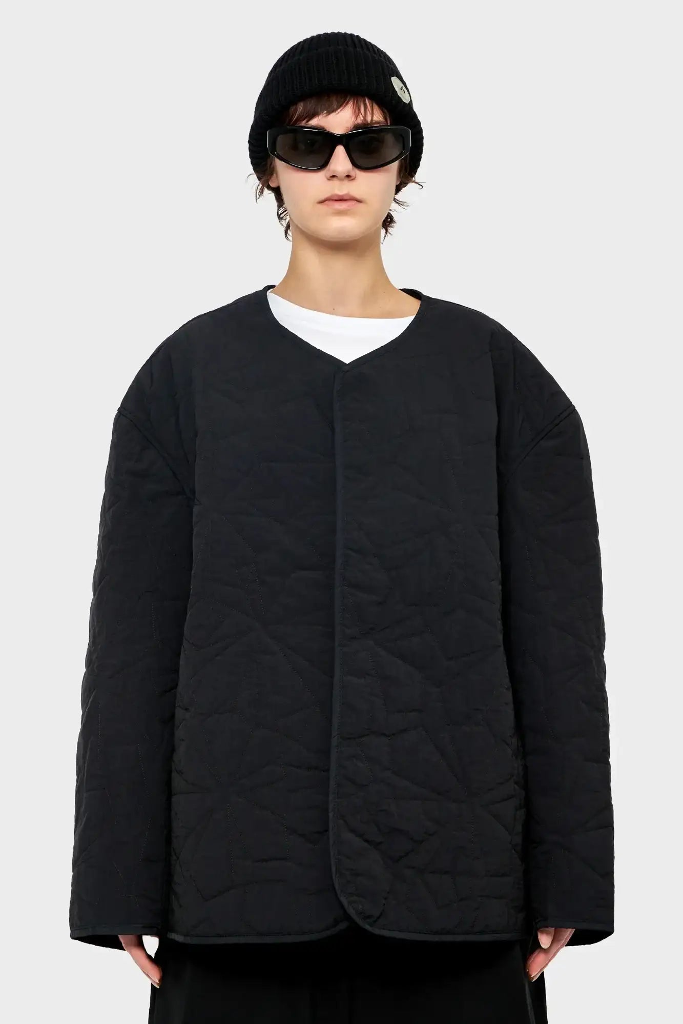 Osoba w czarnej pikowanej kurtce, czapce i okularach przeciwsłonecznych, styl streetwear.