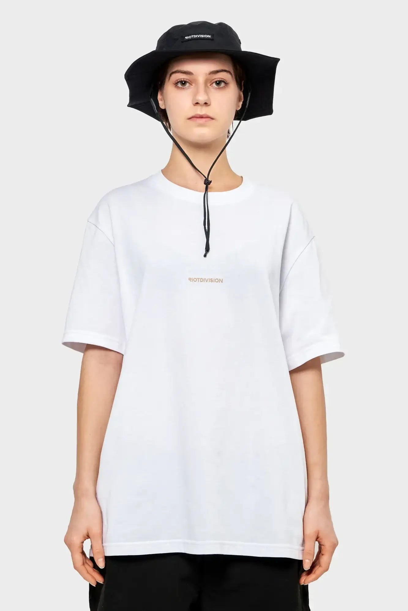 osoba w białym t-shircie RIOTDIVISION i czarnym kapeluszu bucket hat, na jasnym tle