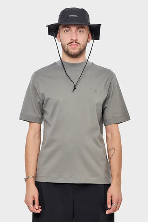 Mężczyzna w szarej koszulce i czarnym kapeluszu z logo RIOTDIVISION, minimalistyczny styl streetwear.