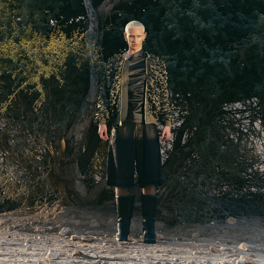 Model w militarnej kurtce, czarnej koszuli i bermudach, stylizowany na tle natury