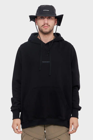 Mężczyzna w czarnej bluzie z kapturem Riotdivision i czarnym kapeluszu typu bucket hat, styl streetwear.