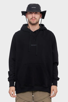 Mężczyzna w czarnej bluzie z kapturem Riotdivision i czarnym kapeluszu typu bucket hat, styl streetwear.