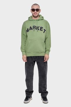 Mężczyzna w zielonej bluzie z kapturem MARKET, czarne jeansy, sneakersy, okulary przeciwsłoneczne