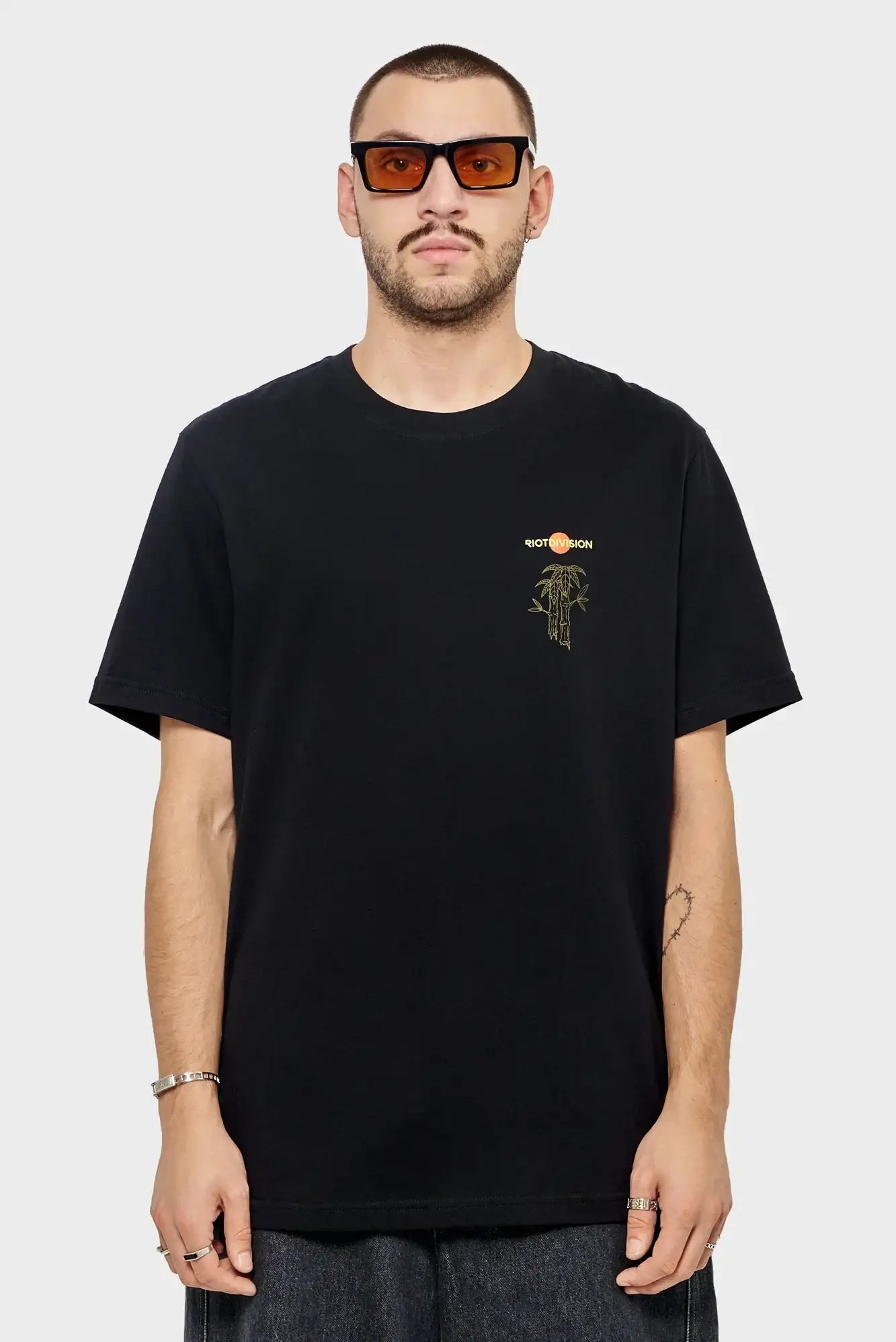 Mężczyzna w czarnej koszulce RIOTDIVISION, pomarańczowe okulary przeciwsłoneczne, styl streetwear