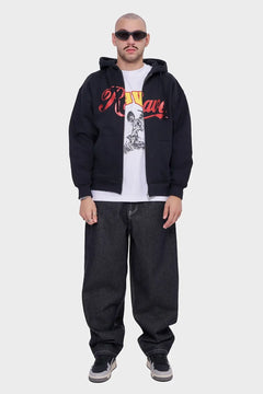 Mężczyzna w czarnej rozpinanej bluzie z kapturem i napisem, białej koszulce z grafiką, szerokich jeansach, sneakersach i okularach przeciwsłonecznych, styl streetwear