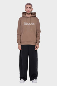 Mężczyzna w brązowej bluzie z kapturem Études, czarne spodnie, białe buty, styl streetwear