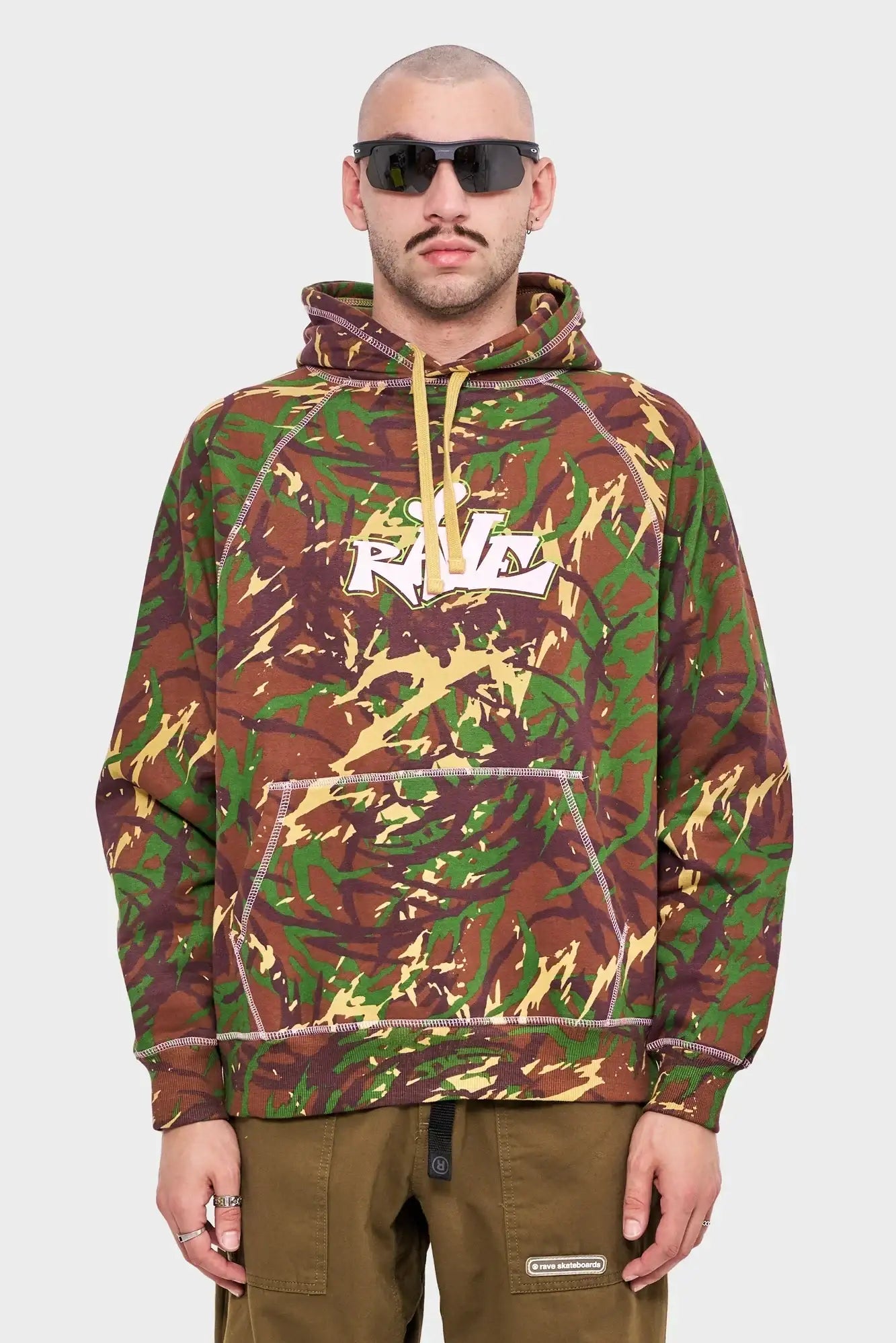 Mężczyzna w okularach przeciwsłonecznych, bluza z kapturem w brązowo-zielony moro wzór z logo RAVE.