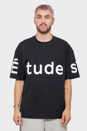 Mężczyzna w czarnej koszulce z dużym białym napisem Études, styl streetwear, tło jasne.