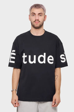 Mężczyzna w czarnej koszulce z dużym białym napisem Études, styl streetwear, tło jasne.