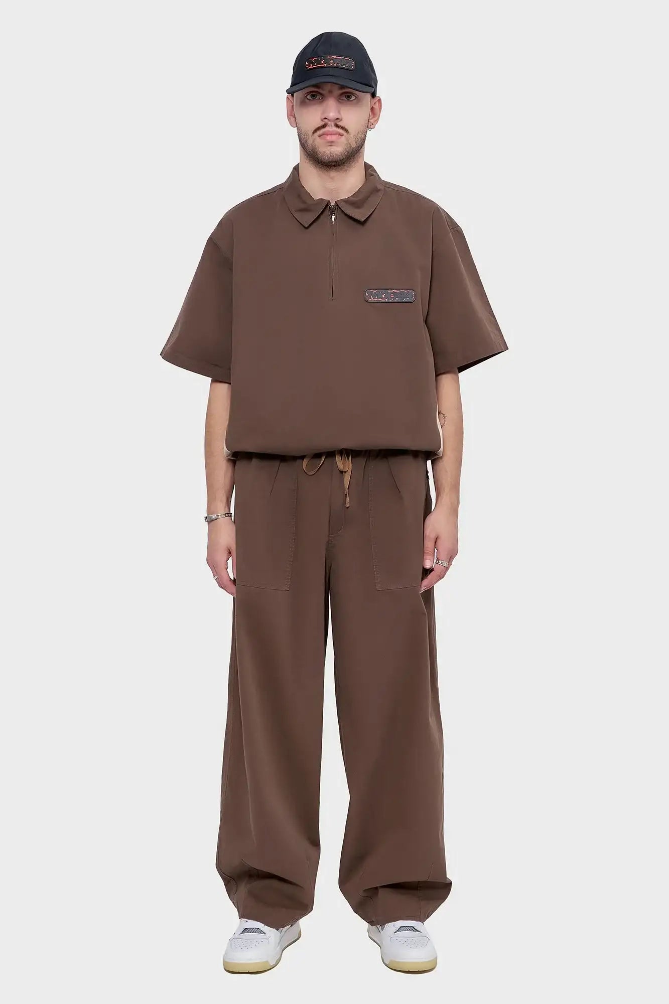 Mężczyzna w brązowym zestawie oversize, spodnie z szerokimi nogawkami, jasne sneakersy, czapka z daszkiem