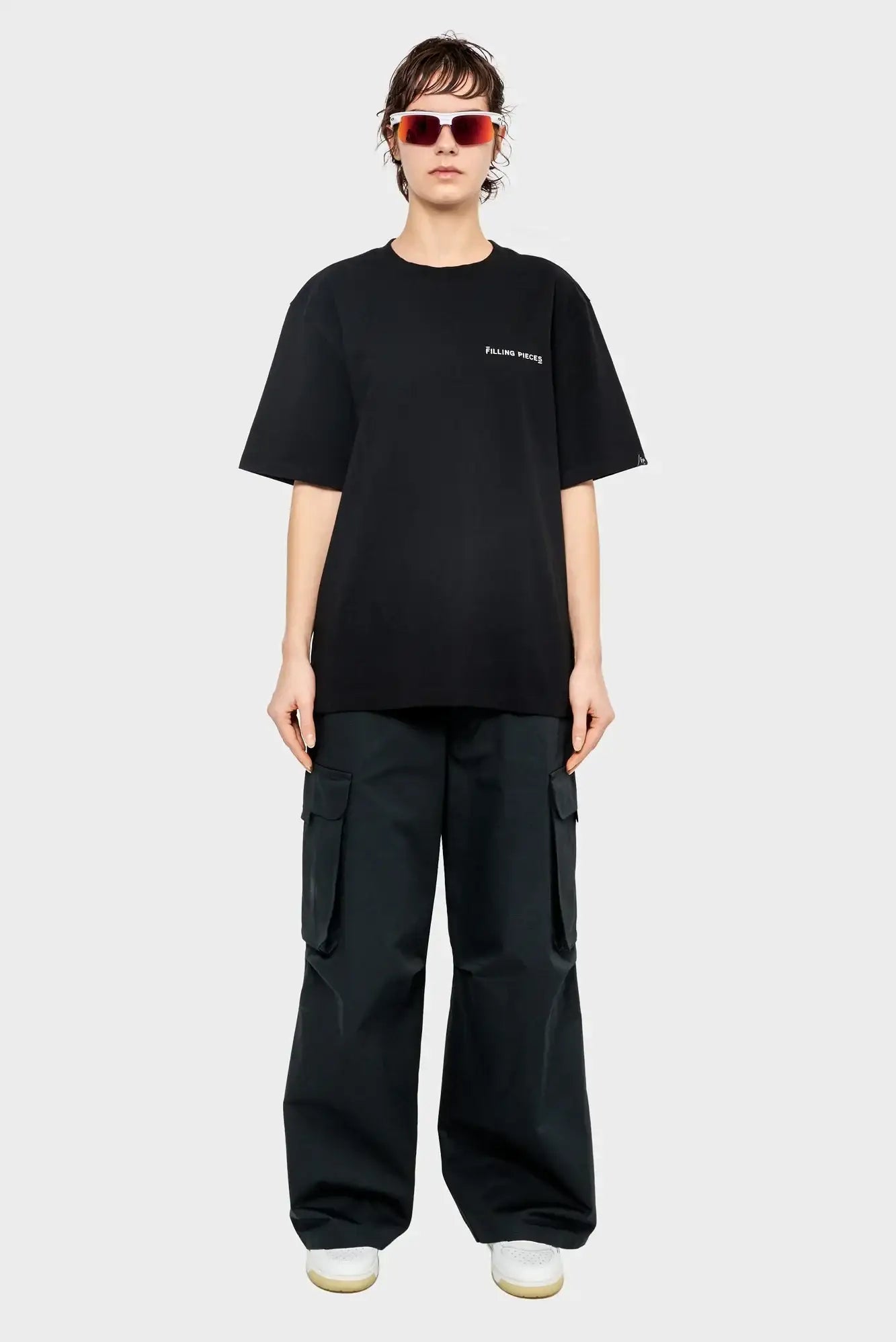 Osoba w okularach przeciwsłonecznych, czarnym t-shircie z napisem, szerokich bojówkach i białych sneakersach, moda streetwear.
