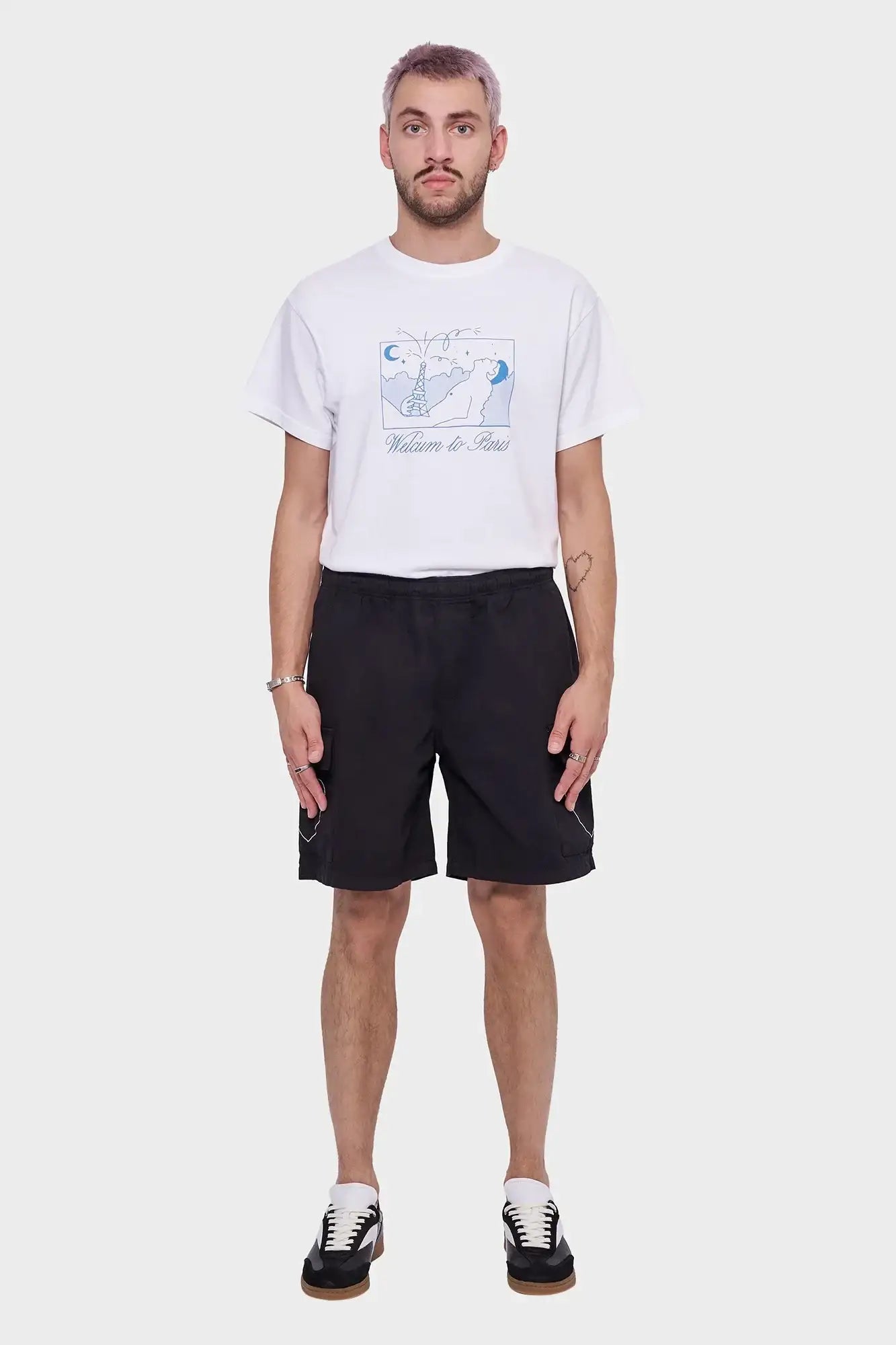Mężczyzna w białej koszulce z nadrukiem Welcome to Paris, czarnych szortach i sneakersach