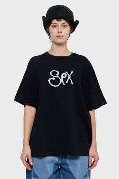 Osoba w czarnej koszulce z napisem, czapka pilotka, jeansy, nowoczesny styl streetwear.