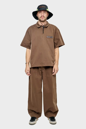 Mężczyzna w brązowym oversize zestawie, szerokie spodnie i koszulka z krótkim rękawem, czarny kapelusz, styl streetwear