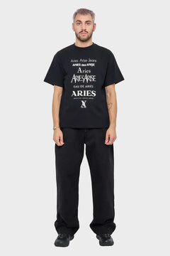 Mężczyzna w czarnej koszulce Aries z białym nadrukiem, czarne spodnie i buty, na jasnym tle