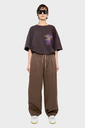 Osoba w ciemnym t-shircie z napisem, szerokich brązowych spodniach i białych sneakersach