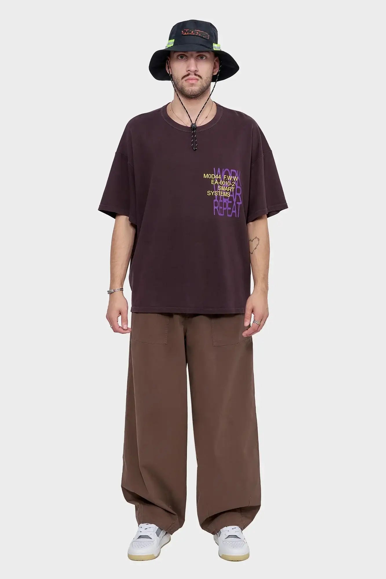 Mężczyzna w brązowym oversized t-shircie z nadrukiem, szerokich spodniach i ciemnym kapeluszu typu bucket hat, styl streetwear