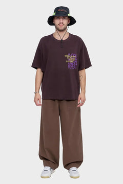 Mężczyzna w brązowym oversized t-shircie z nadrukiem, szerokich spodniach i ciemnym kapeluszu typu bucket hat, styl streetwear