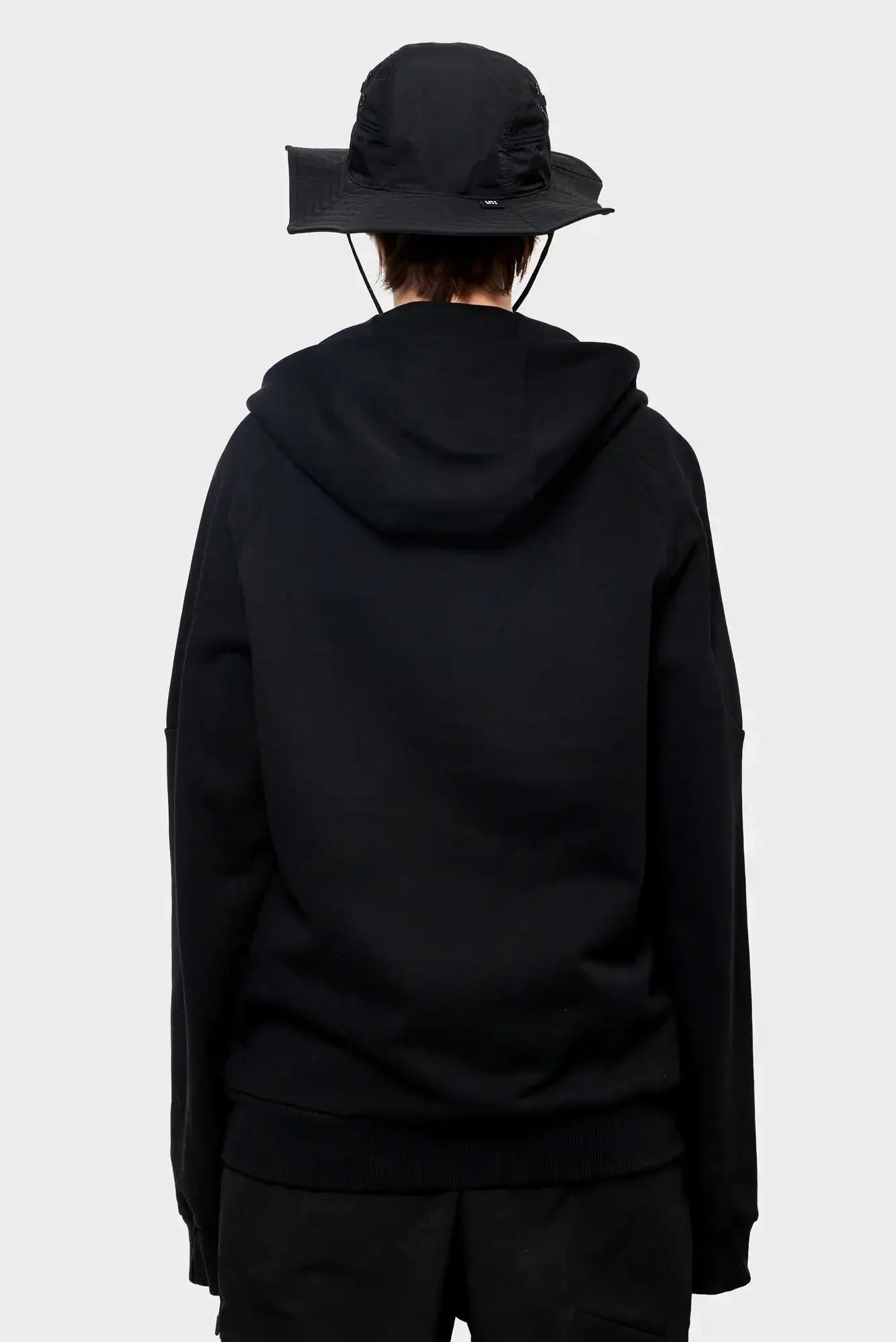 Osoba w czarnej bluzie z kapturem i czarnym kapeluszu typu bucket hat, widok z tyłu.