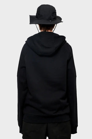 Osoba w czarnej bluzie z kapturem i czarnym kapeluszu typu bucket hat, widok z tyłu.