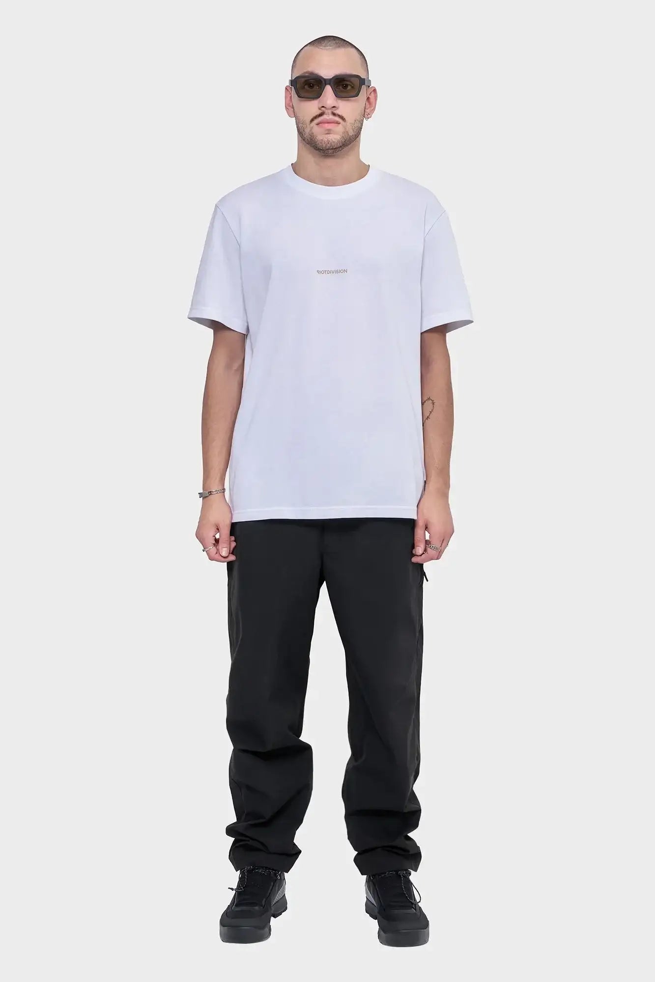 Mężczyzna w białej koszulce ROTDIVISION, czarne spodnie, ciemne okulary, styl streetwear.