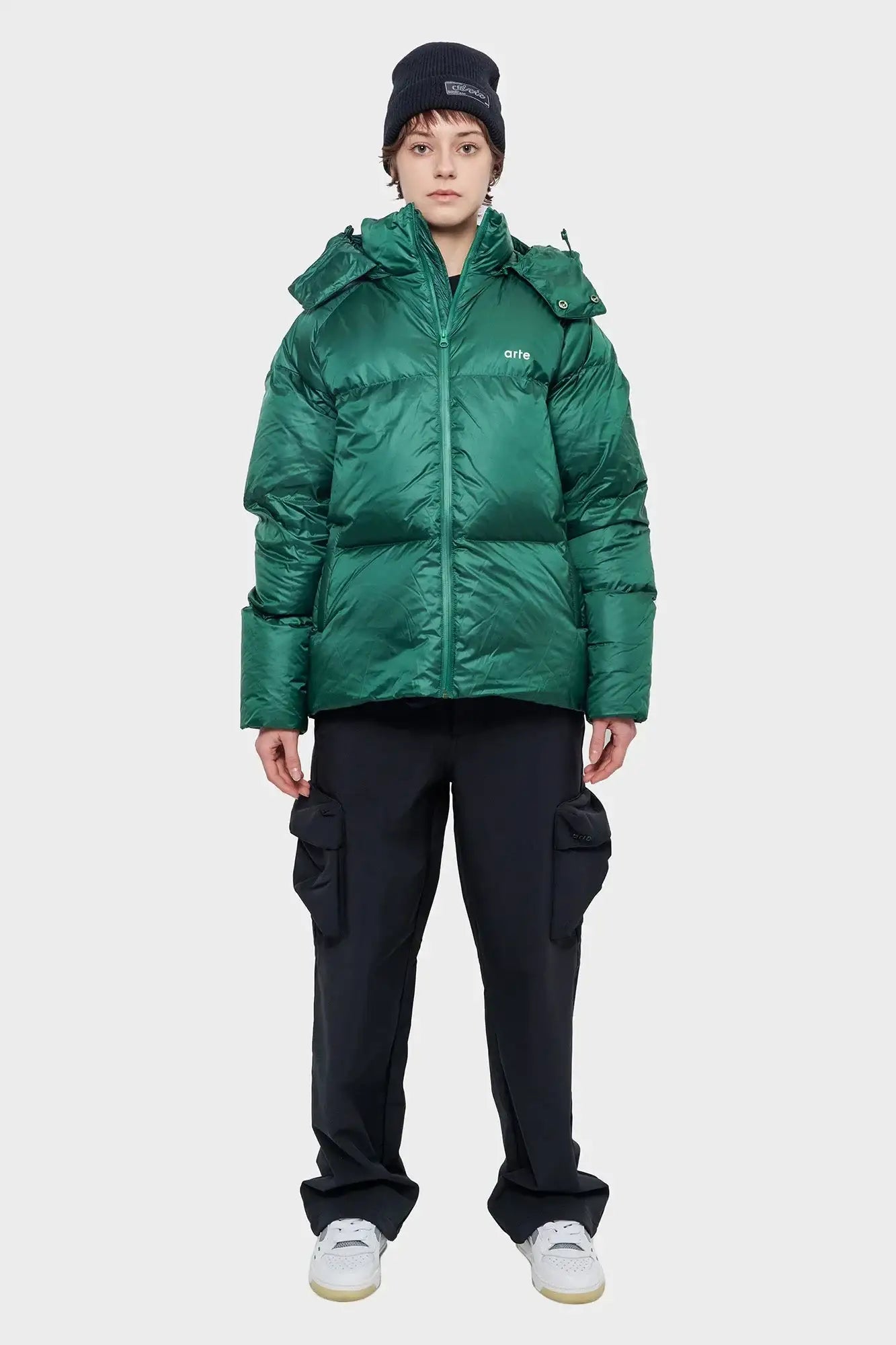 Osoba w zielonej puchowej kurtce Arte, czarnej czapce i czarnych spodniach cargo, styl streetwear