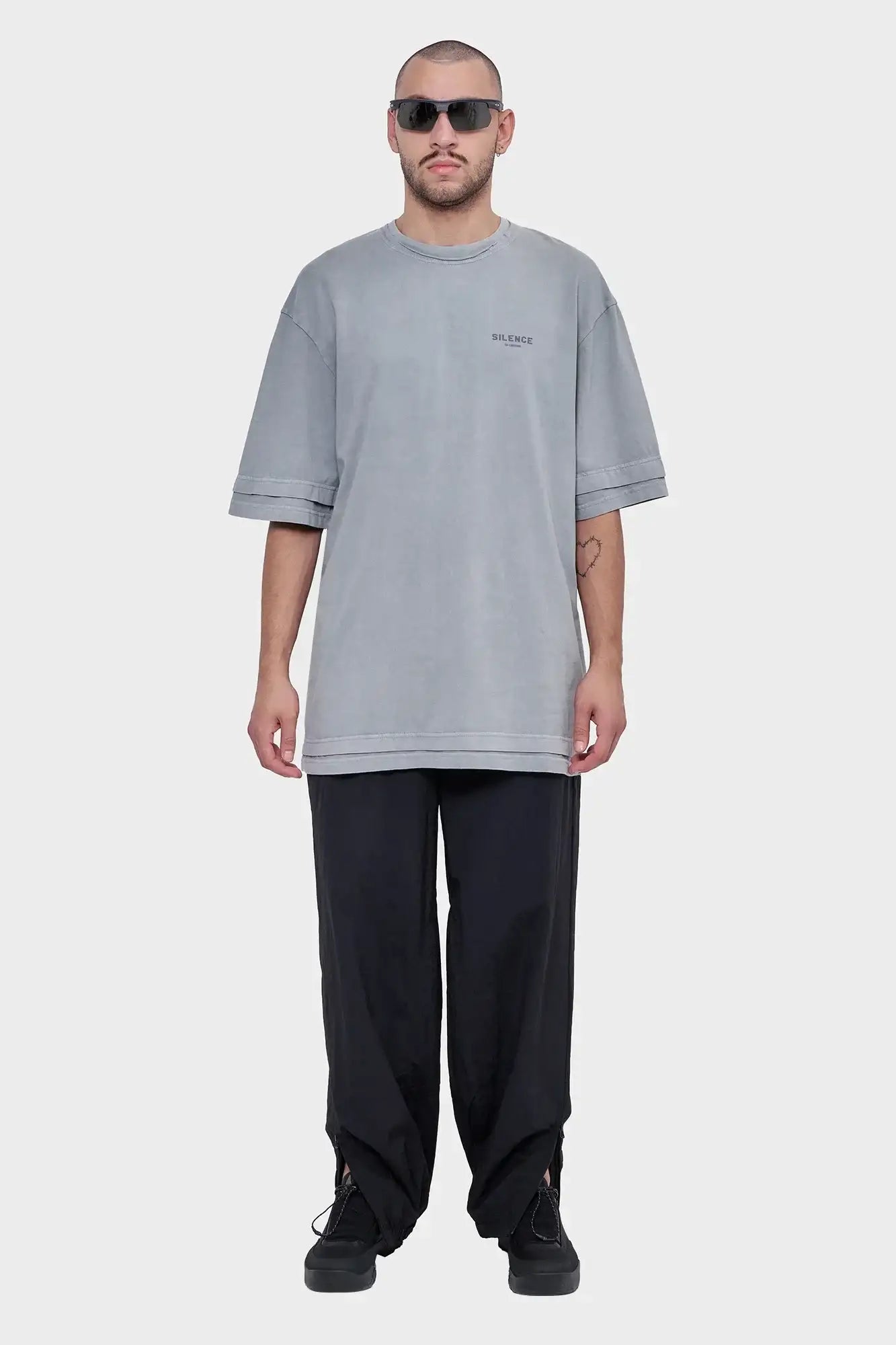 Mężczyzna w szarym oversize t-shircie z napisem SILENCE, czarnych luźnych spodniach i okularach przeciwsłonecznych