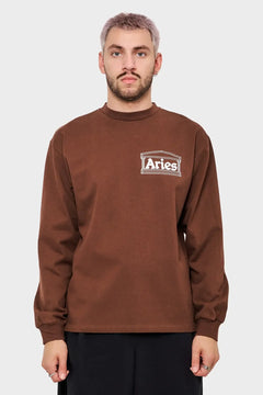 Mężczyzna w brązowej bluzie z długim rękawem i logo Aries na klatce piersiowej.