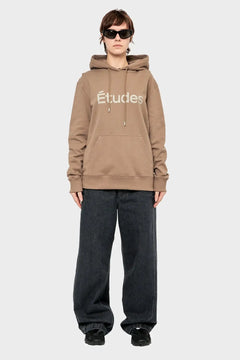 Osoba w brązowej bluzie z kapturem z napisem Études, szerokie czarne jeansy, styl streetwear.
