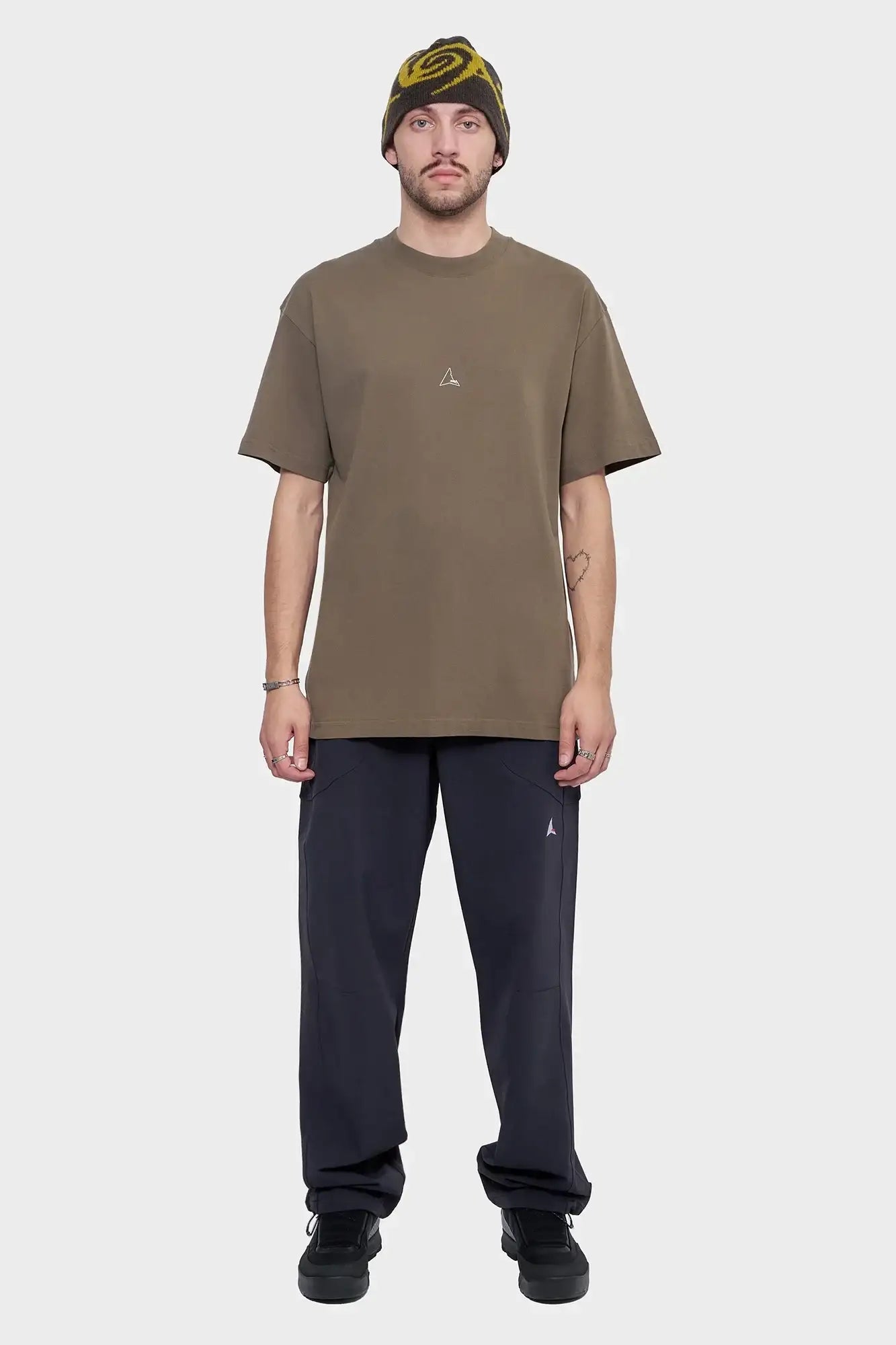 Mężczyzna w brązowym t-shircie, ciemnych spodniach i czapce z żółtym wzorem, styl streetwear