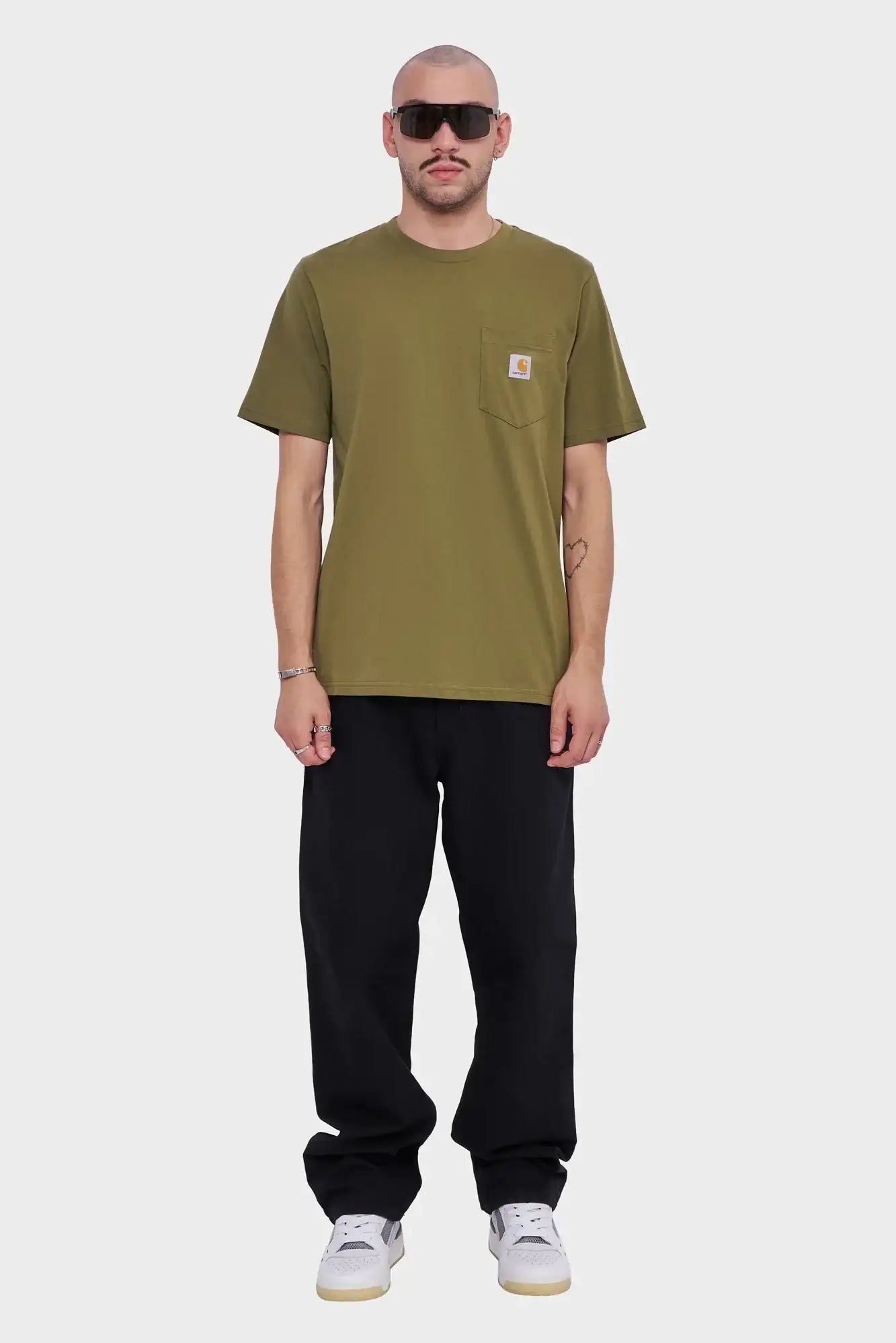 Mężczyzna w oliwkowym t-shircie z kieszonką, czarnych spodniach i białych sneakersach na szarym tle.