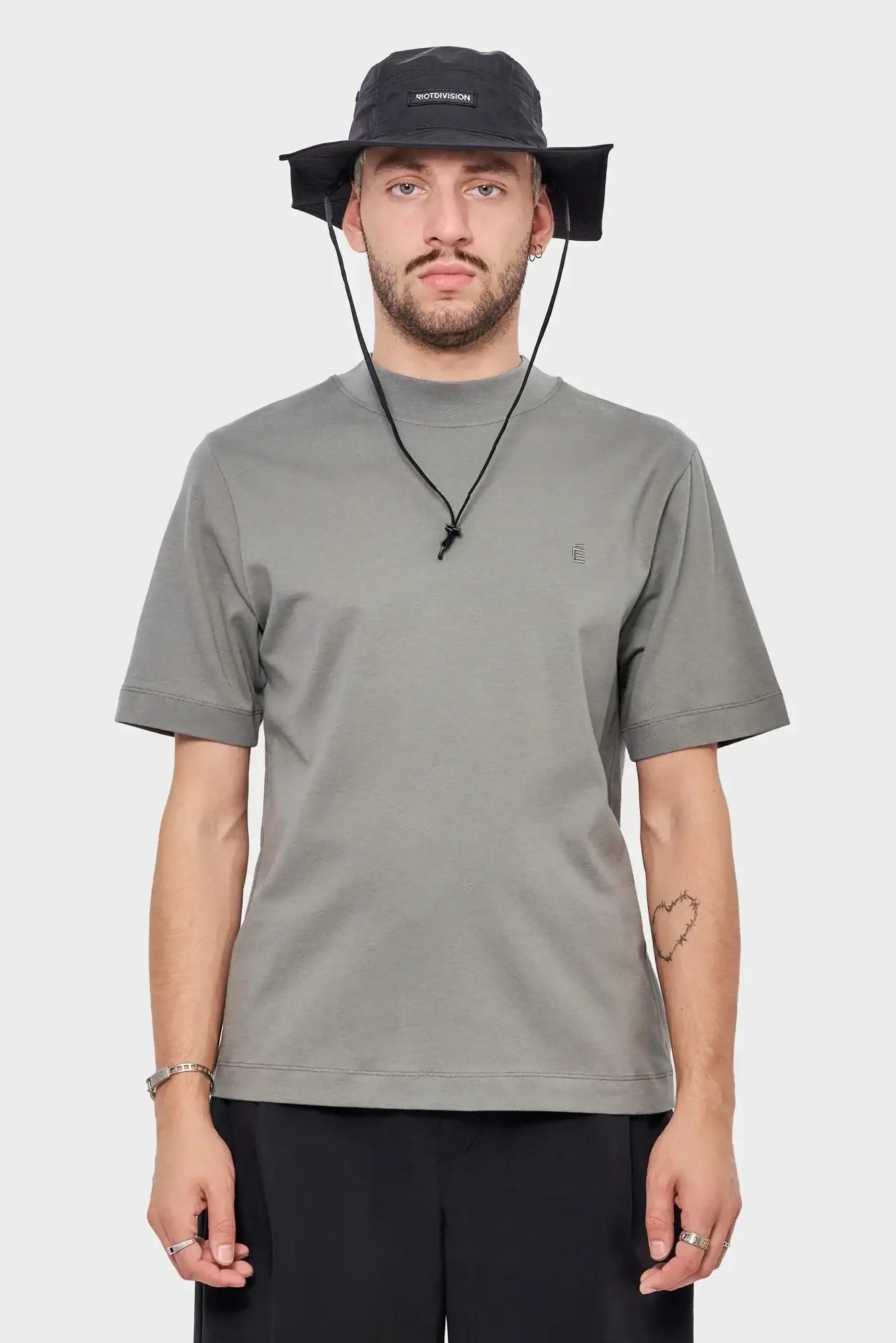 Mężczyzna w szarej koszulce i czarnym kapeluszu z logo RIOTDIVISION, minimalistyczny styl streetwear.