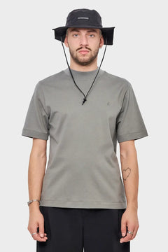 Mężczyzna w szarej koszulce i czarnym kapeluszu z logo RIOTDIVISION, minimalistyczny styl streetwear.