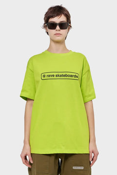 Modelka w limonkowej koszulce rave skateboards, brązowe spodnie, okulary przeciwsłoneczne