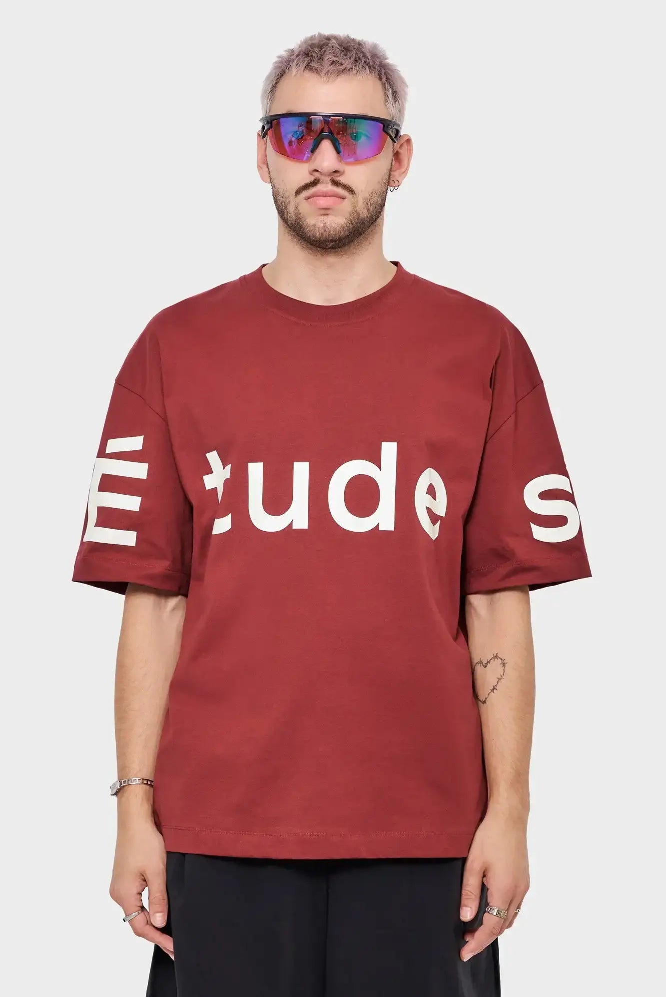 Mężczyzna w bordowej koszulce ÉTUDES, sportowe okulary, minimalistyczny styl streetwear