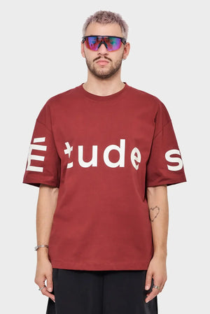 Mężczyzna w bordowej koszulce ÉTUDES, sportowe okulary, minimalistyczny styl streetwear