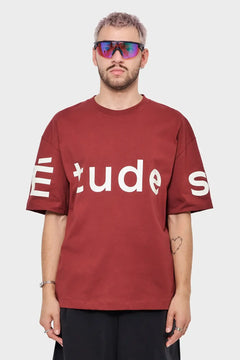 Mężczyzna w bordowej koszulce ÉTUDES, sportowe okulary, minimalistyczny styl streetwear