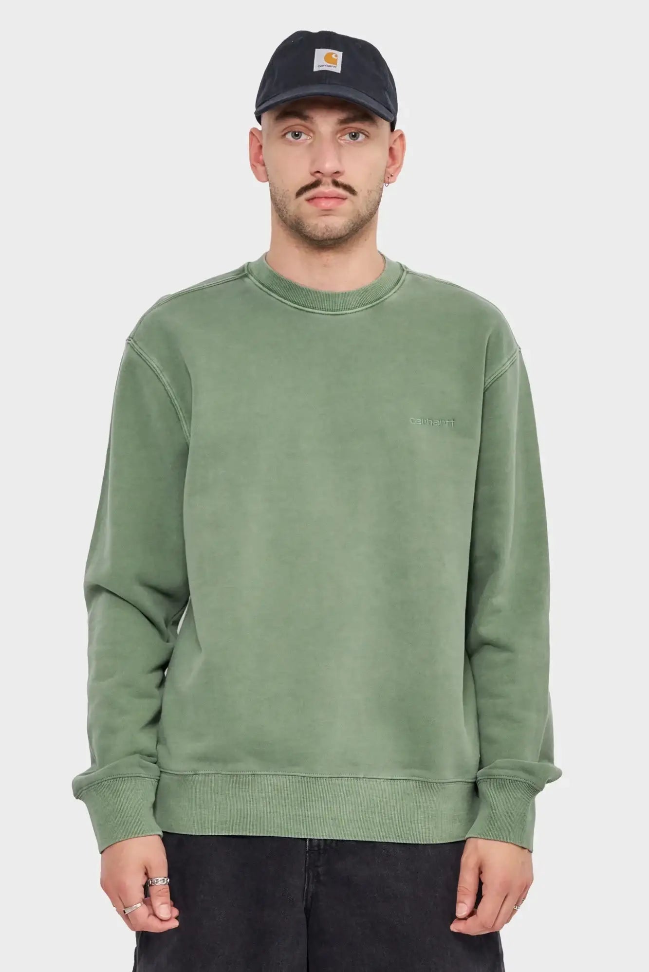 Mężczyzna w zielonej bluzie Carhartt WIP i czapce z logo, neutralne tło, odzież streetwear