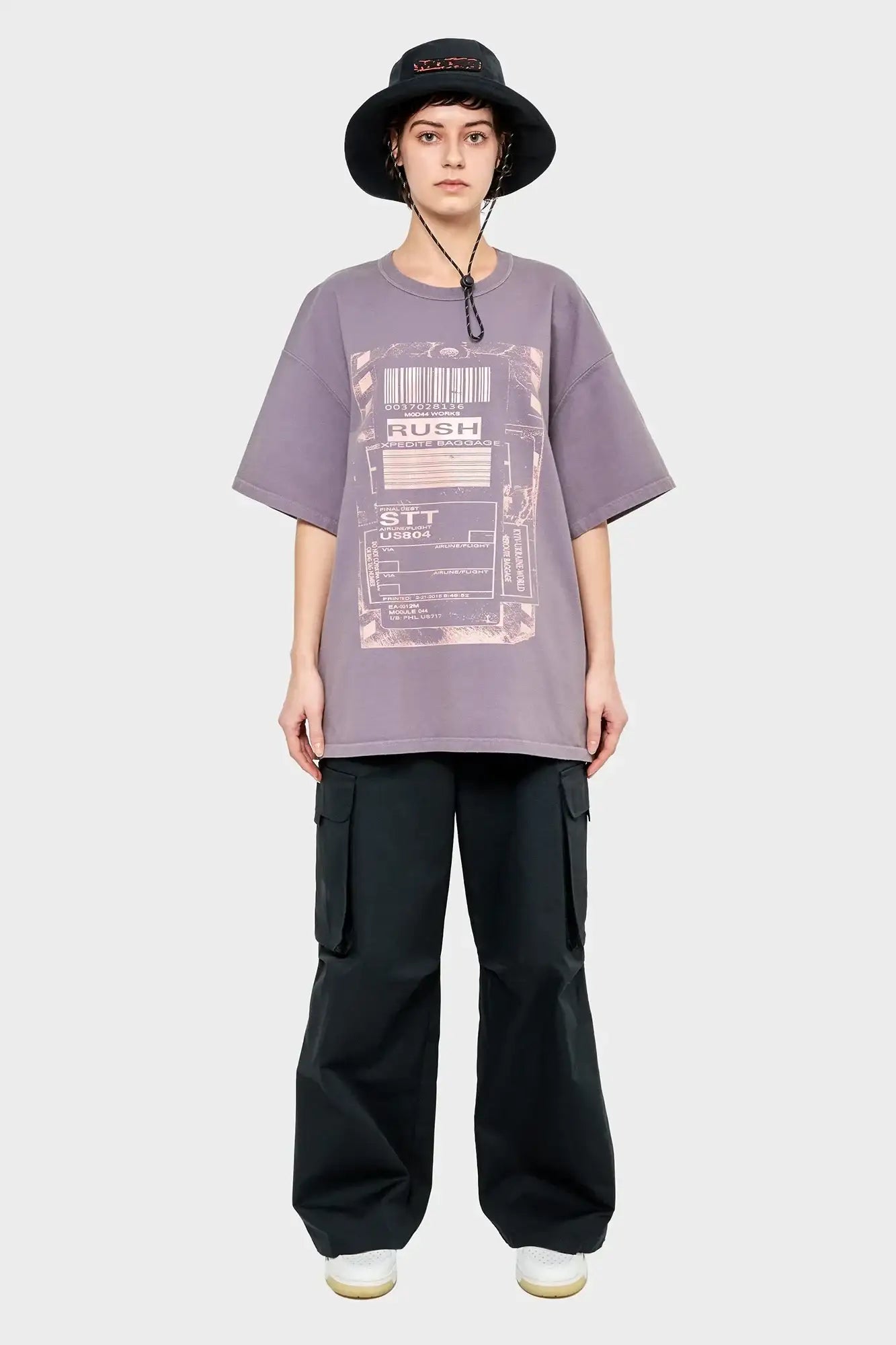 Osoba w luźnym fioletowym t-shircie z nadrukiem, czarnych bojówkach i czarnym kapeluszu typu bucket hat