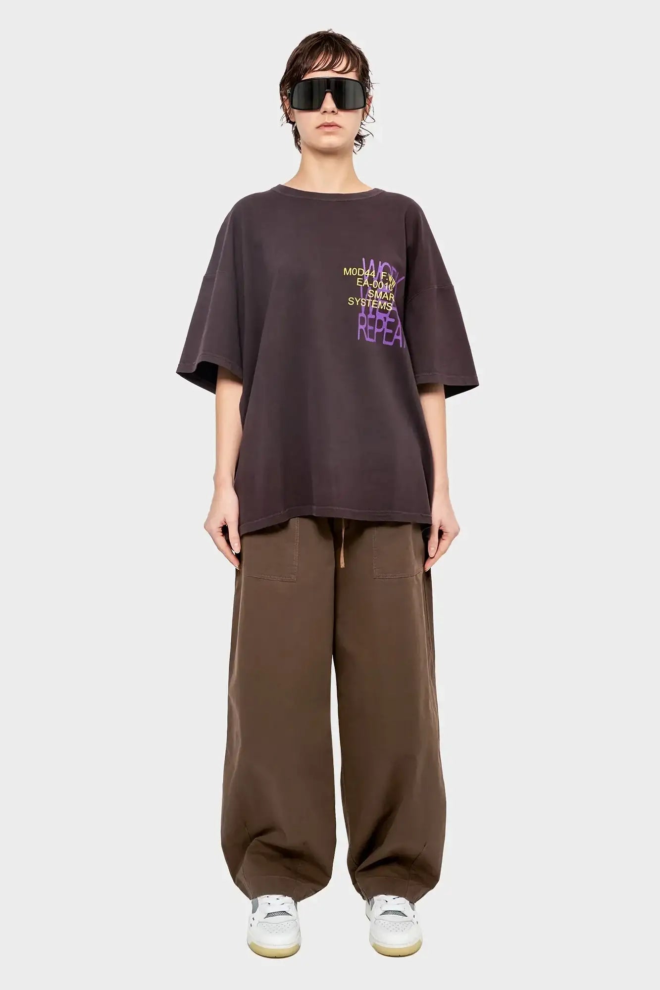 Modelka w oversize'owej ciemnobrązowej koszulce z nadrukiem i szerokich brązowych spodniach, modne okulary, styl streetwear.