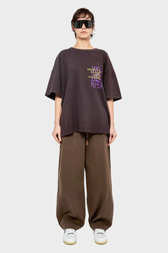 Modelka w oversize'owej ciemnobrązowej koszulce z nadrukiem i szerokich brązowych spodniach, modne okulary, styl streetwear.