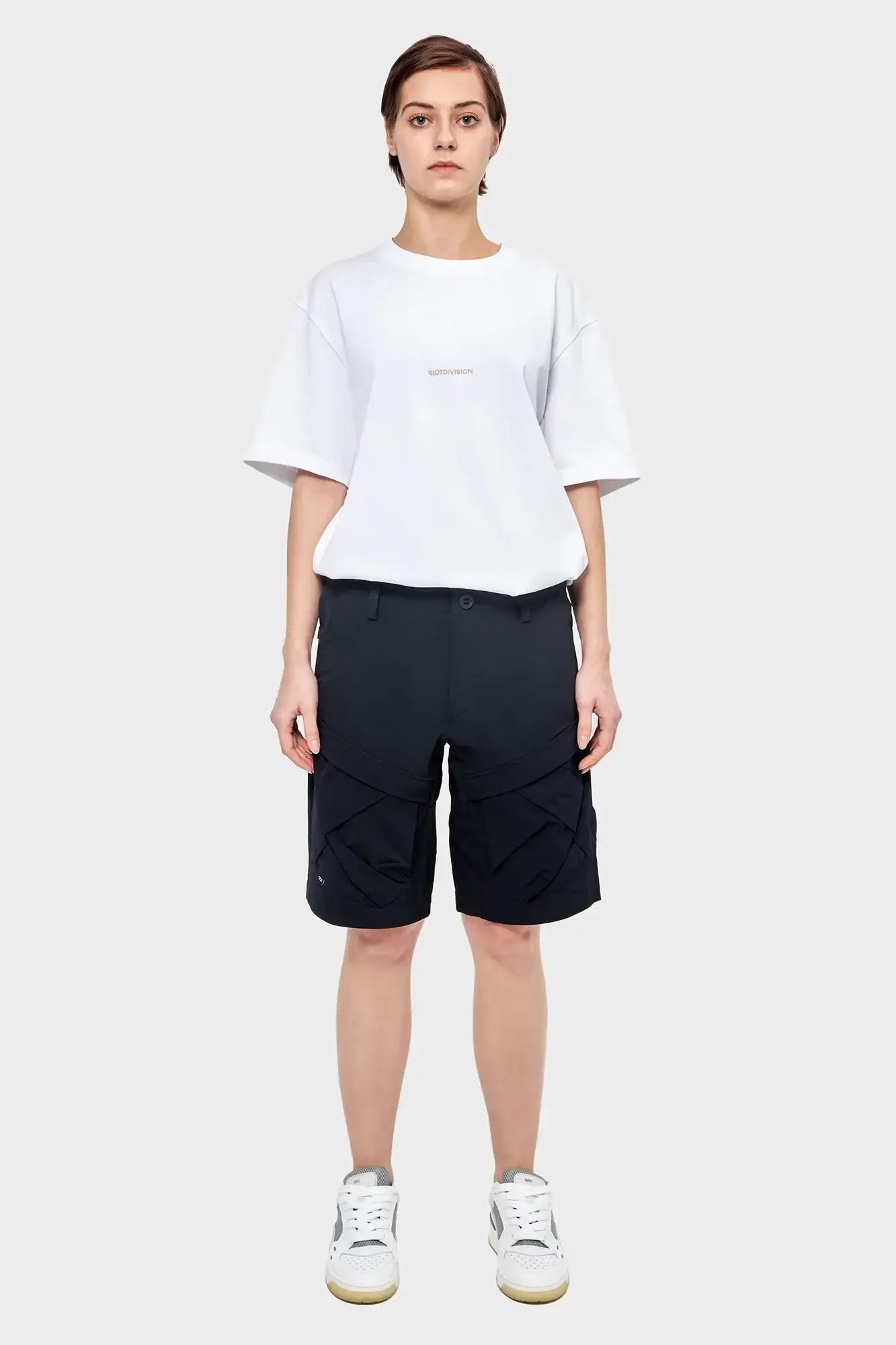 Modelka w białym t-shircie RIOTDIVISION, czarnych szortach i białych sneakersach na szarym tle