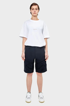 Modelka w białym t-shircie RIOTDIVISION, czarnych szortach i białych sneakersach na szarym tle