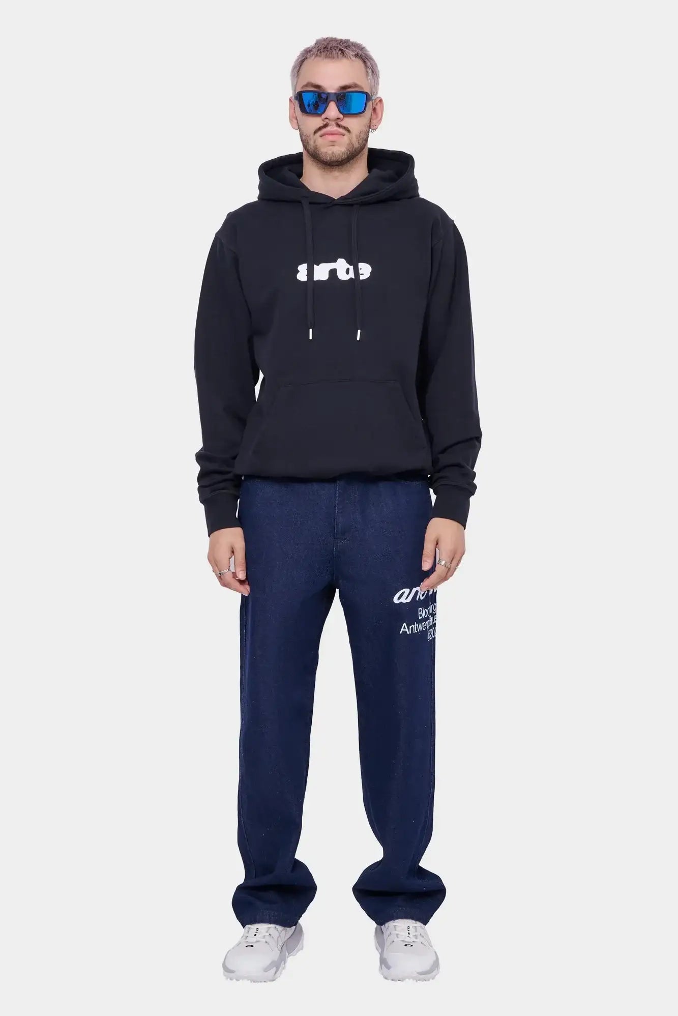 Mężczyzna w czarnej bluzie z kapturem z białym logo, granatowych jeansach i białych sneakersach, styl streetwear.