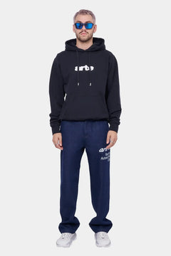 Mężczyzna w czarnej bluzie z kapturem z białym logo, granatowych jeansach i białych sneakersach, styl streetwear.