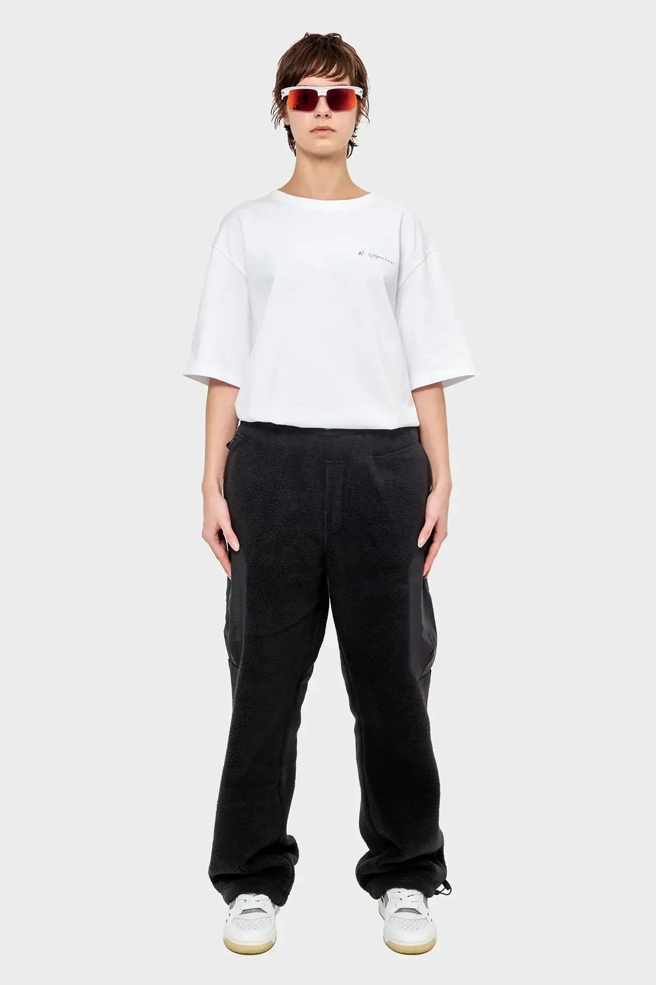 Osoba w białym t-shircie, czarnych spodniach dresowych, białych sneakersach i okularach przeciwsłonecznych, styl streetwear.